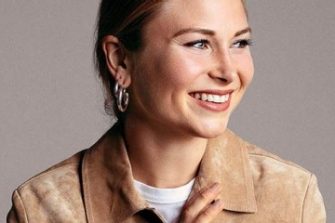 Grace Tame smiling and looking to her left, with her hair tied up and wearing a camel coloured shirt
