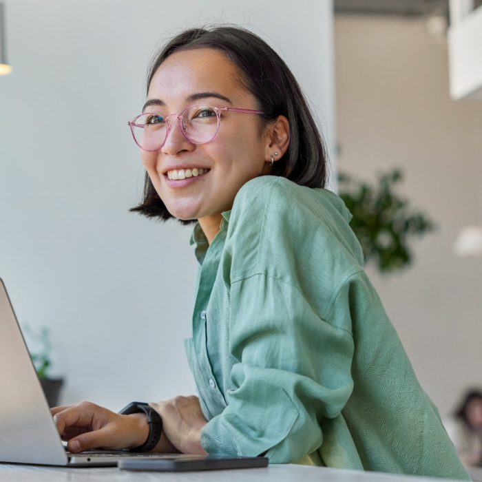 Pretty cheerful Asian woman in eyeglasses and casual clothes browses laptop computer connected to 4g internet updates software uses modern technologies poses in cafeteria looks gladfully away