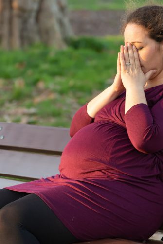 Young pregnant woman taking a moment to de-stress and relax as she suffers morning sickness seated in warm sunshine on a park bench with her hands covering her face and eyes closed