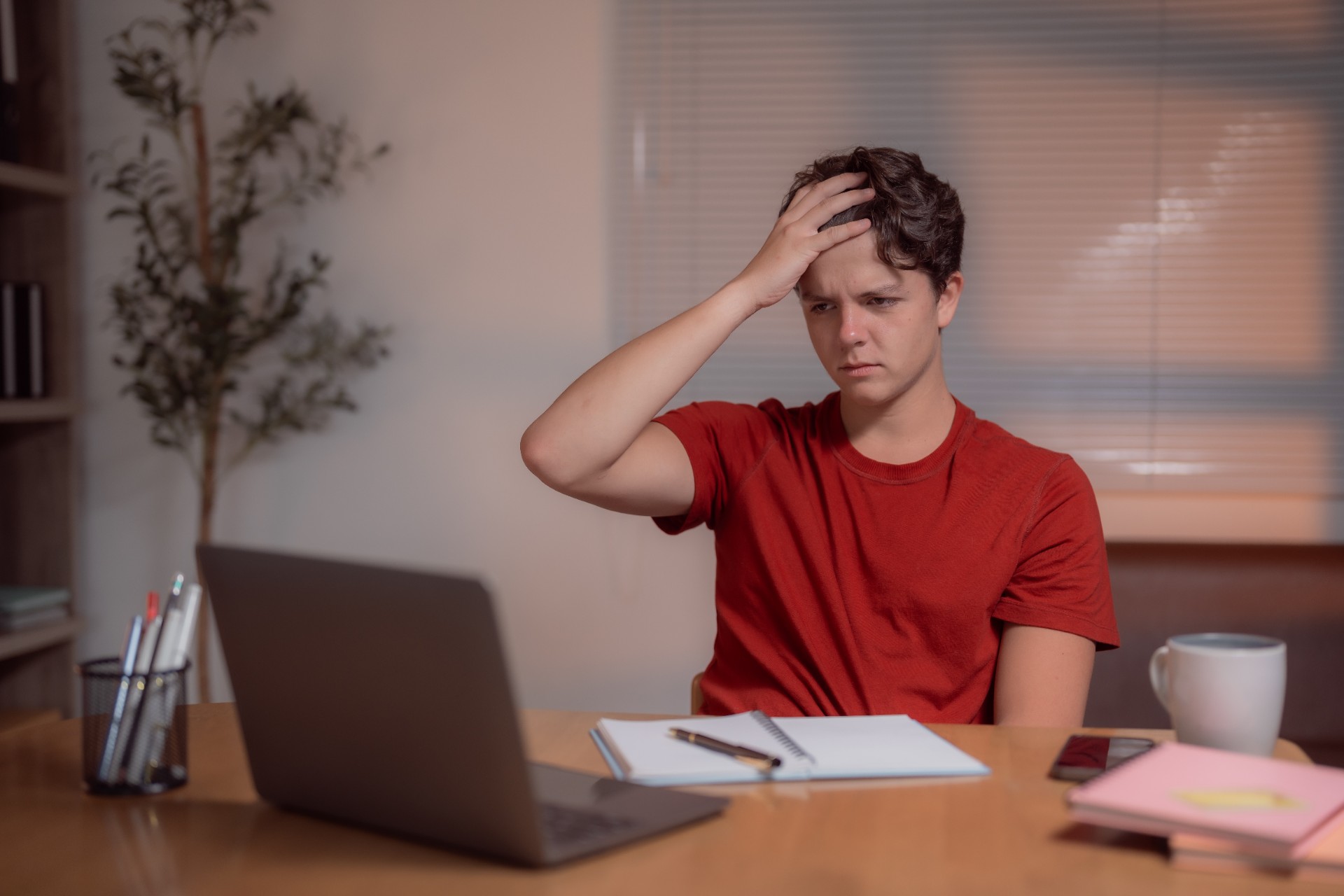 Young man touching his head with his hand while looking at his laptop, experiencing frustration and headache during a late-night study session at his home desk