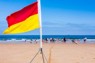 A red and yellow lifeguard safety flag on a beach with families and children in the background playing in the sea