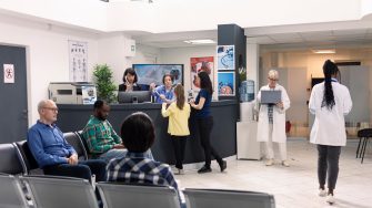 Busy hospital lobby with patients and visitors seated in waiting area. Receptionist and nurse assist at front desk as diverse individuals prepare for medical appointments and consultations.