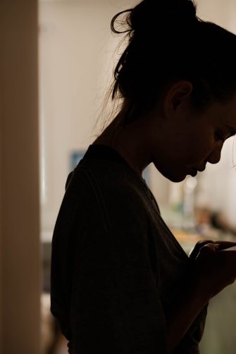 Teenager in a black hoodie with a hood on is sitting on a bench, covering face with her hands, back view