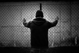 A black and white shot from behind of a young male detainee  in front of a fence