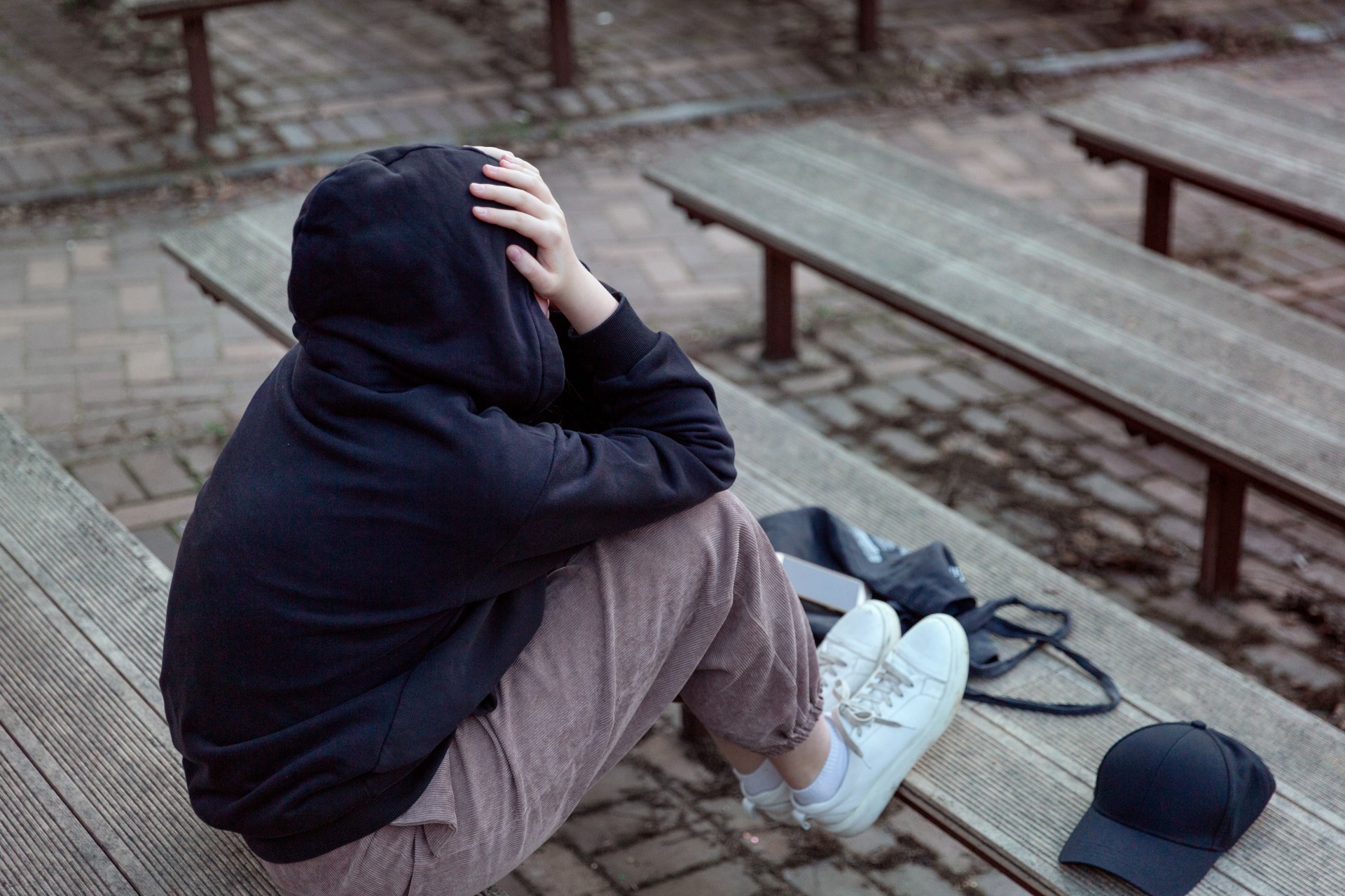 Teenager in a black hoodie with a hood on is sitting on a bench, covering her face with her hands, back view