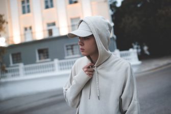 Young teenage boy 18-19 year old wear trendy beige hoodie and cap work out over city background early in morning in dark. Selective focus. Teenagerhood. Active lifestyle. Sad teenager have bad mood. 
