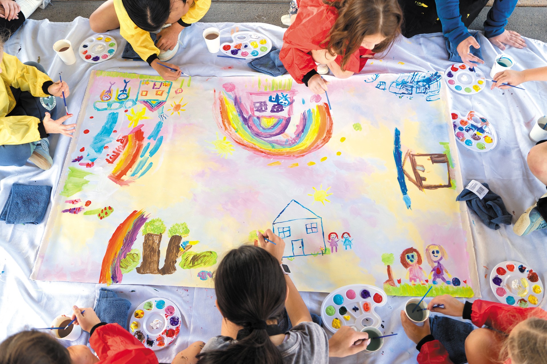 Children sit in a circle around a large piece of fabric painting homes, and rainbows and family