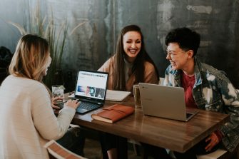 Three women laughing while sitting at a table with laptops