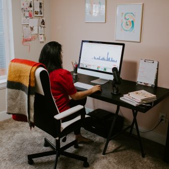 a woman sits at her home workspace, looking at a computer screen 