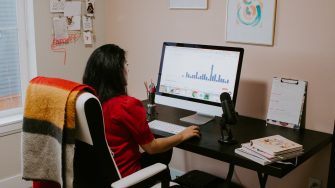 a woman sits at her home workspace, looking at a computer screen 