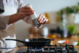An anonymous chef with apron standing at the gas stove and pouring salt into frypan.