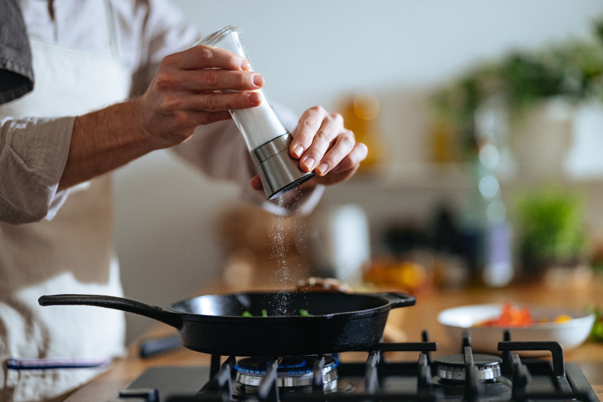 An anonymous chef with apron standing at the gas stove and pouring salt into frypan.