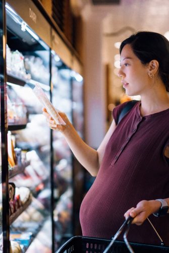 Young Asian pregnant woman grocery shopping in supermarket, choosing fresh packets of cheese from the diary aisle. Eating well with balanced nutrition. Pregnancy health and wellness. Healthy eating habit and lifestyle during pregnancy
