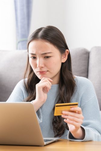 A young woman sits in front of her latop holding a credit card and looking frustrated.
