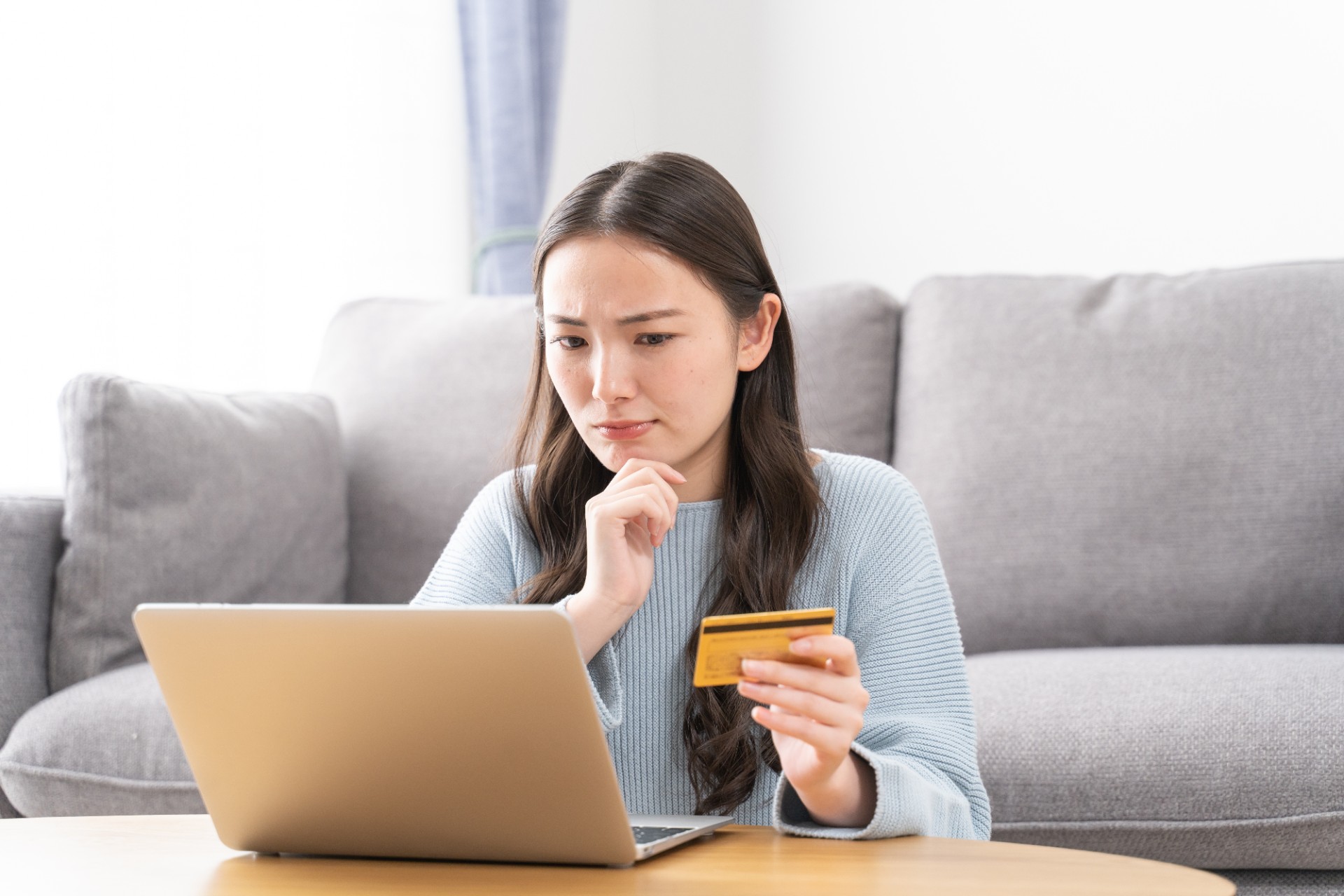 A young woman sits in front of her latop holding a credit card and looking frustrated.