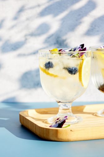 Chilled cocktails with fresh citrus fruits garnished with edible flowers on a wooden serving board. Summer drinks with soda and fruit.