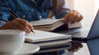 Casual young female student handwriting information on diary notebook while reading book and work on laptop computer with white cup of coffee on the table at home.