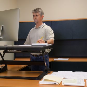 A man is working at a standup desk in an office where he works because standing is healthier than sitting all day. Live healthy, don’t sit all day.
