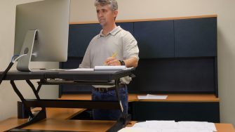 A man is working at a standup desk in an office where he works because standing is healthier than sitting all day. Live healthy, don’t sit all day.