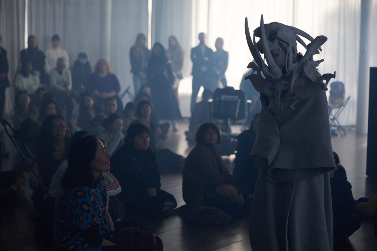 A dark room with spectators looking at a robed figure with a bone headress