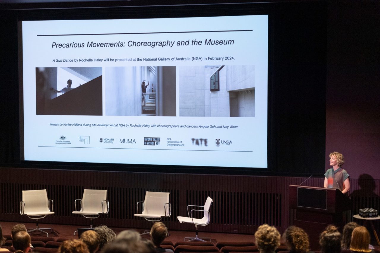 A person speaks at a lectern with a screen