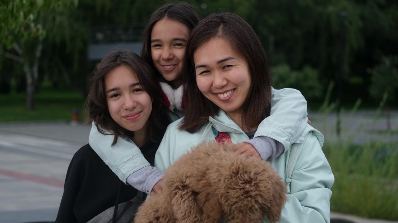 Smiling beautiful mixed race family, mom, little brown poodle and two daughters, posing for the camera in a city park.