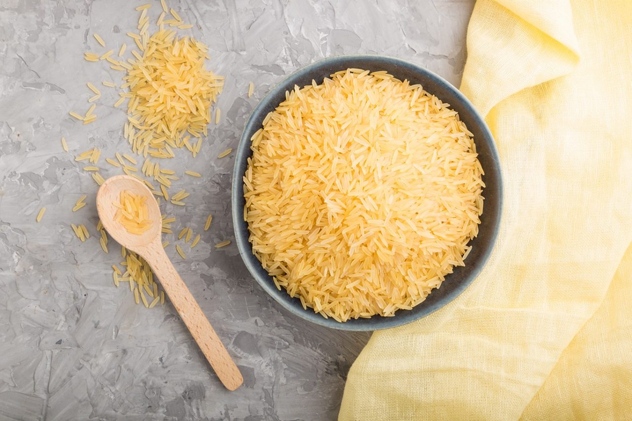 Blue ceramic bowl with raw golden rice and wooden spoon on a gray concrete background and yellow textile. Top view, flat lay, close up.